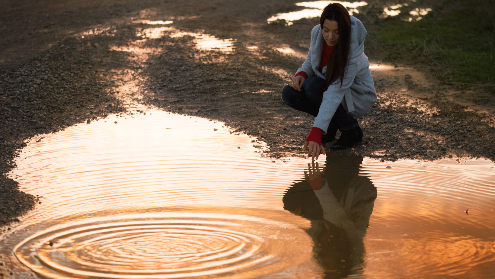 Person crouching beside a puddle on a dirt road, touching the water and creating ripples.