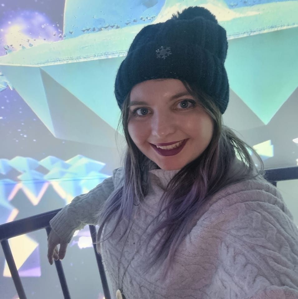 Katie is a white female, smiling wearing a blue knitted hat with brown and purple hair and a grey knitted jumper. She has purple lipstick and is standing infront of a projector screen that has blue icebergs.