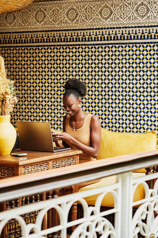 A woman sitting in front of a laptop in a lounge area while texting on her cellphone.