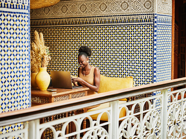 A woman sitting in front of a laptop in a lounge area while texting on her cellphone.