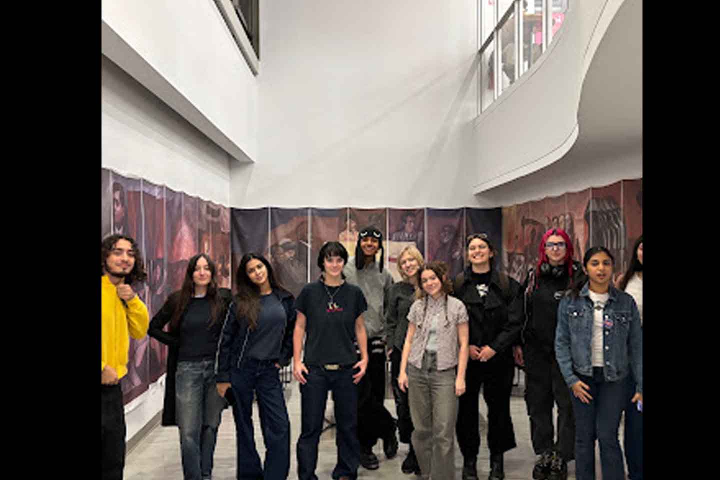 A group of students stand in a space. Behind them, occupying the back and side walls, hangs a reproduction of a mural by José Clemente Orozco.