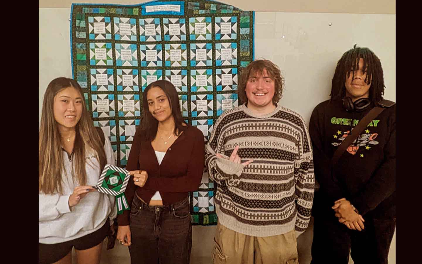 Four students in front of a quilt with squares pertaining to elements of the day's debate. 