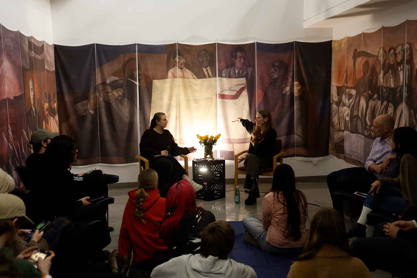 Photo of people listening to Alhena Katsof talk with Emily Clayton in a space with an Orozco mural reproduced on cloth drapes hanging from the walls.