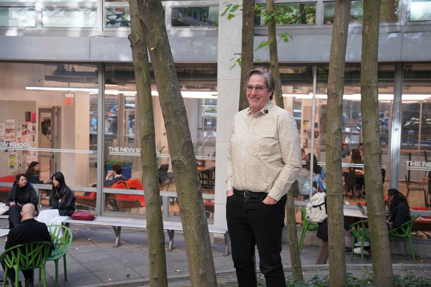Photo of Preston Prince standing among some birch trees in the Lang courtyard. Behind him are people sitting in the courtyard and in the Lang Cafe where glass windows look out onto the courtyard.