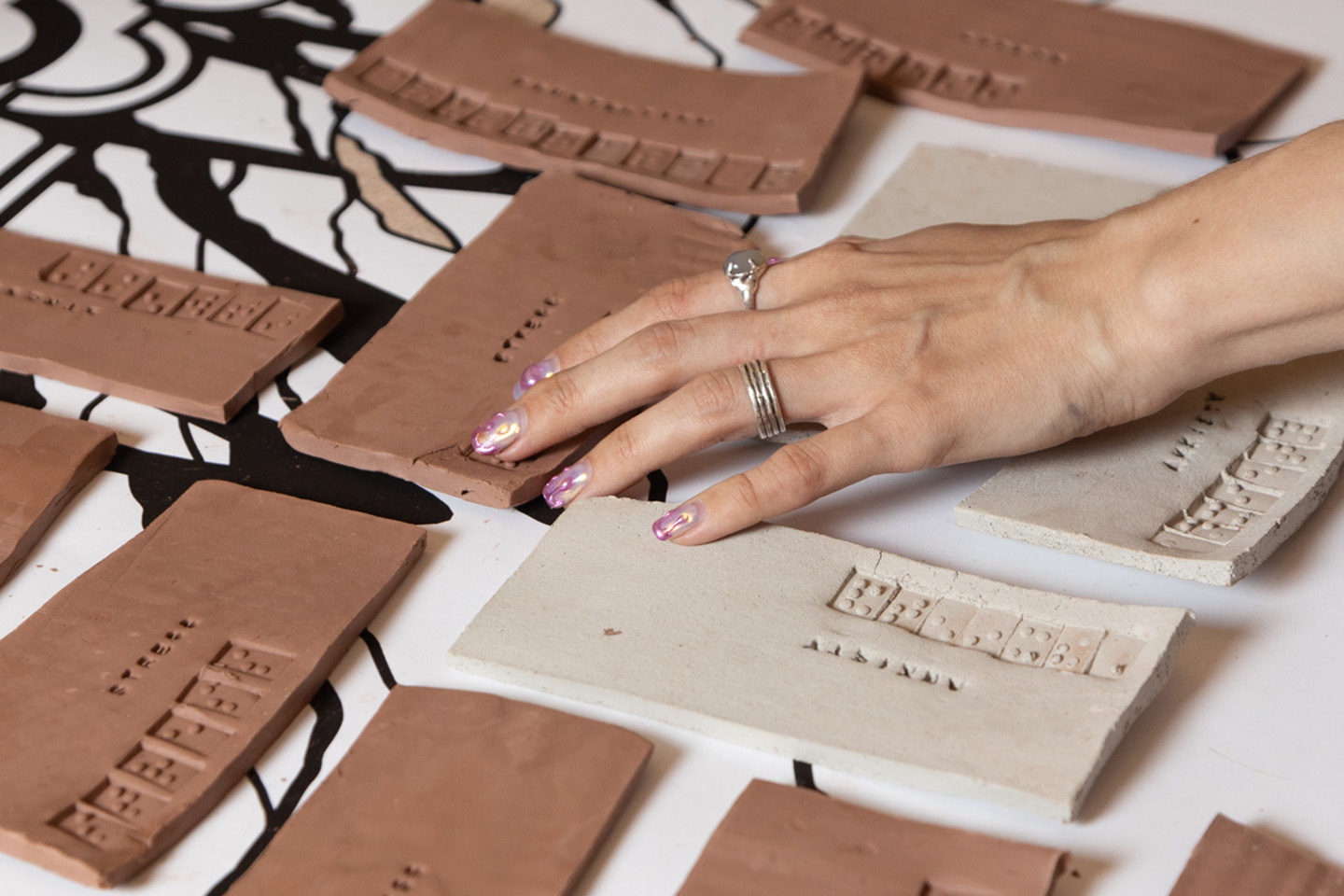 Close-up view of a student’s hand touching thick, neutral colored ceramic like tiles laid out across a table. Custom clay tiles are stamped with braille.