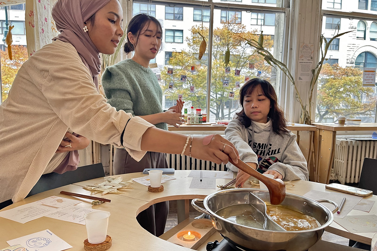 Students sit around a uniquely designed community table with one student serving broth as part of an exploration in food, ritual, and materiality.