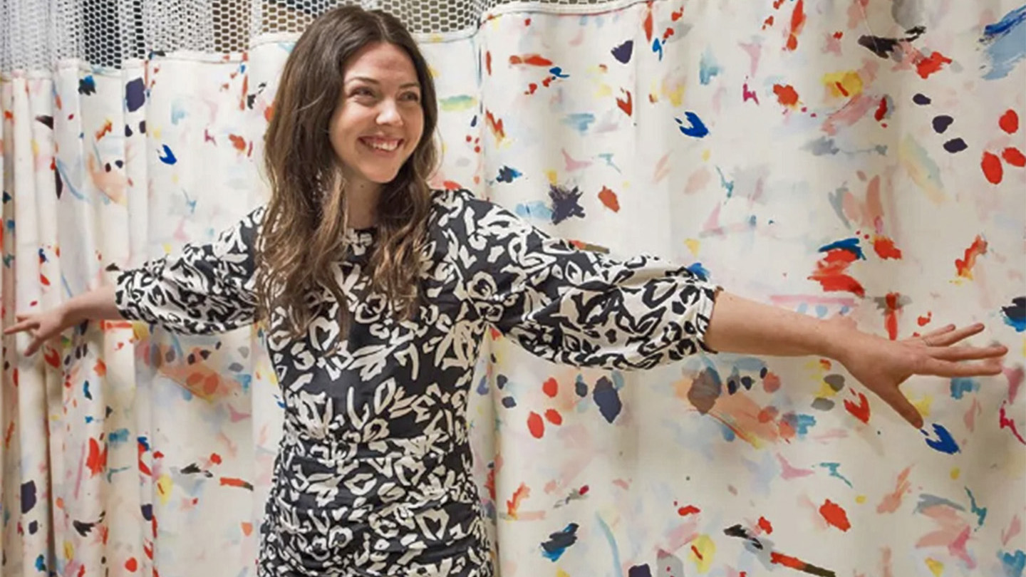 Tanya Taylor, smiling, standing with arms akimbo in front of a curtain with colorful spatters on it.