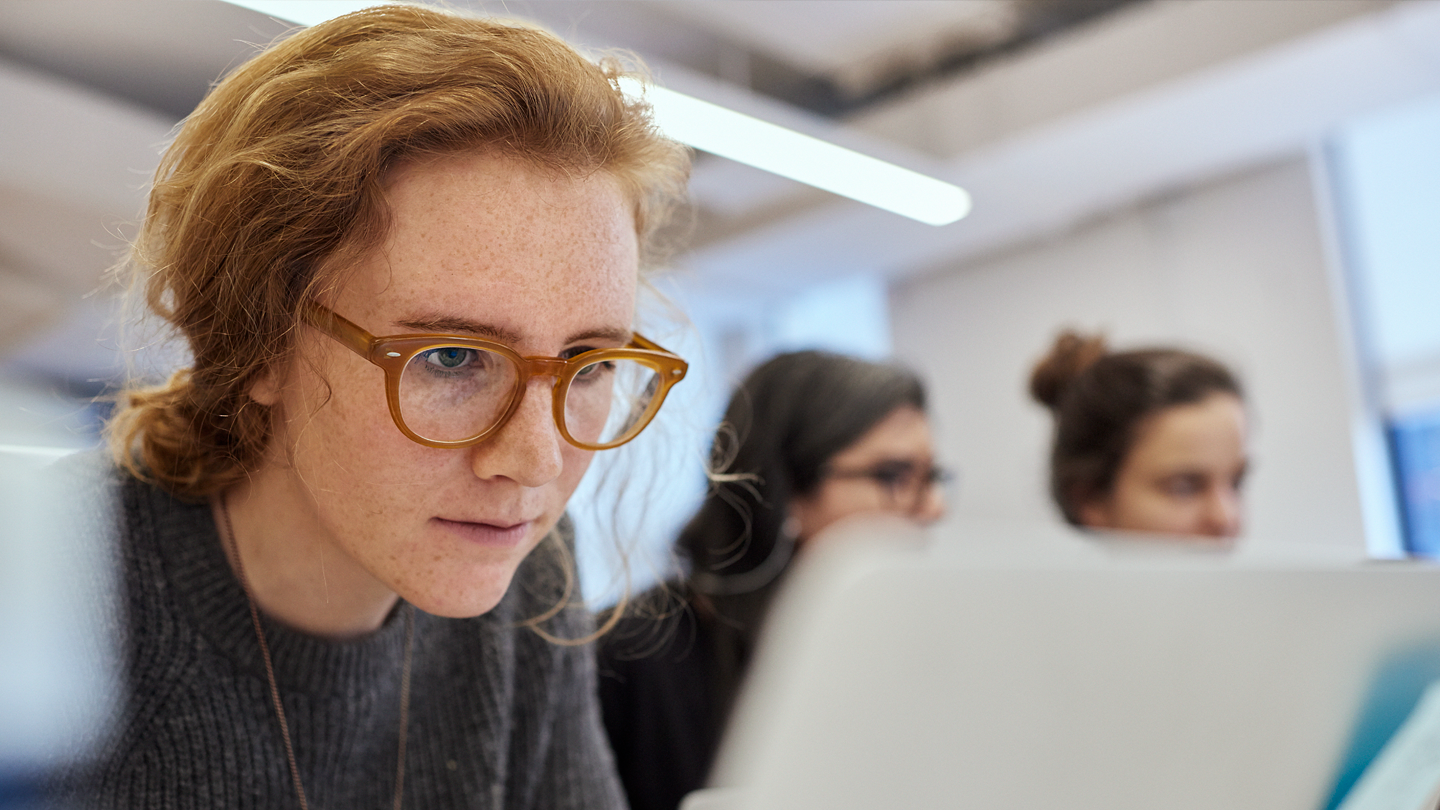 A student with red hair and brown glasses intently viewing a laptop near two out of focus students in the background.