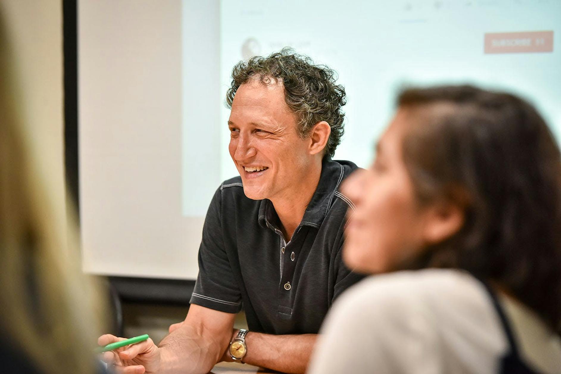 A professor smiles at students in a classroom with a dropdown screen behind.