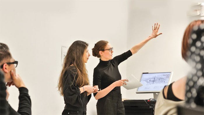 A standing professor demonstrates something with their arm up. A student stands next to her; others are seated. Everyone is wearing black.