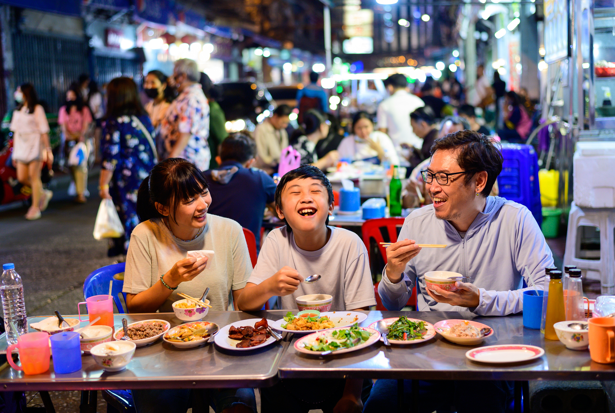 A man, woman, and child smiling as they eat street food with a crowd behind them in Bangkok, Thailand