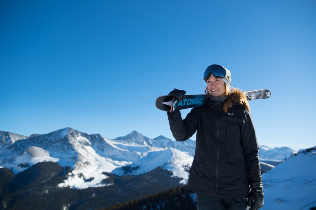Allie Welsh poses with her skis at the top of Copper Mountain with peaks in the background. She's trying to make it to the Olympics in halfpipe skiing.