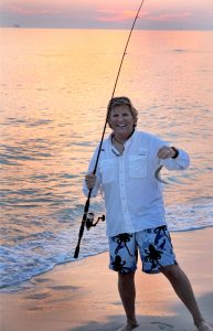 A man shows a fish he caught from the beach. The sun sets in the background.