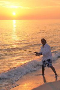 As the sun sets, man fishes from a beach.