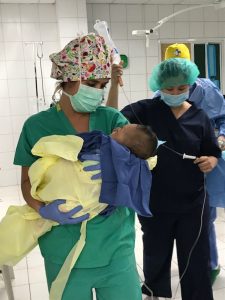 Nurse Drea Mogenson holds a baby in her arms in the Philippines. She is wearing her surgical mask and scrubs while working for a Colorado nonprofit called Mending Faces.