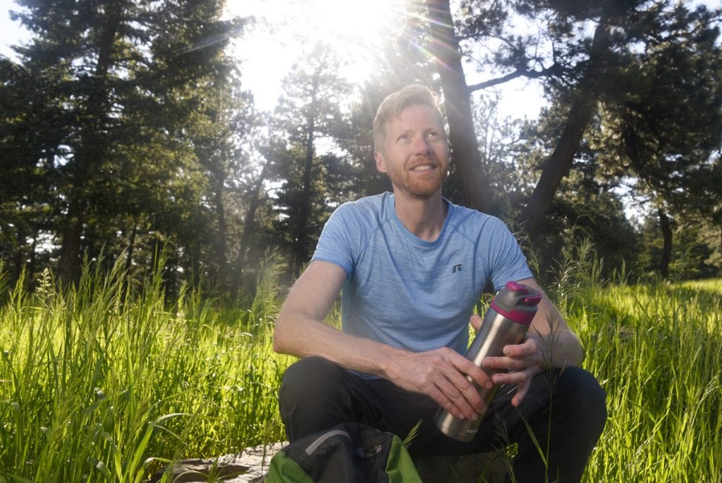 Jonathan Proctor, an environmentalist and former wilderness ranger, sits on a rock in the forest. The sun is bright behind him. He was born with a condition called pectus excavatum. His breast fone was pressing in on his heart and causing shortness of breath.