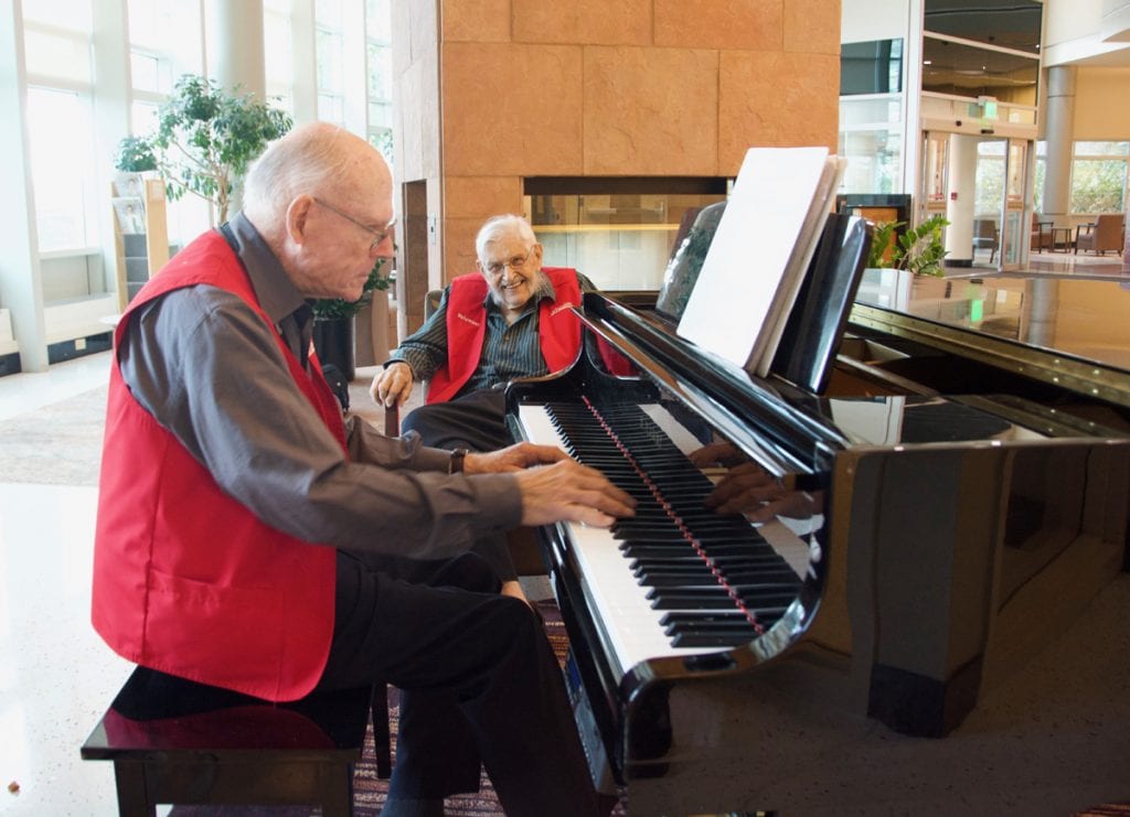 Two older men in red volunteer vests perform at UCHealth University of Colorado Hospital. One is 81 and is playing the piano in the foreground. The other is a 94-year-old WW II vet. He's in the backgrounda and is smiling before he sings.