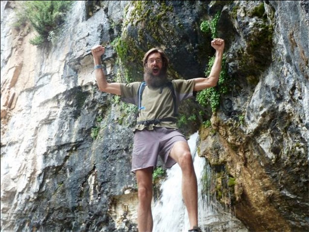 Andy Bonnett holds his arms up while hiking. A waterfall is in the background.