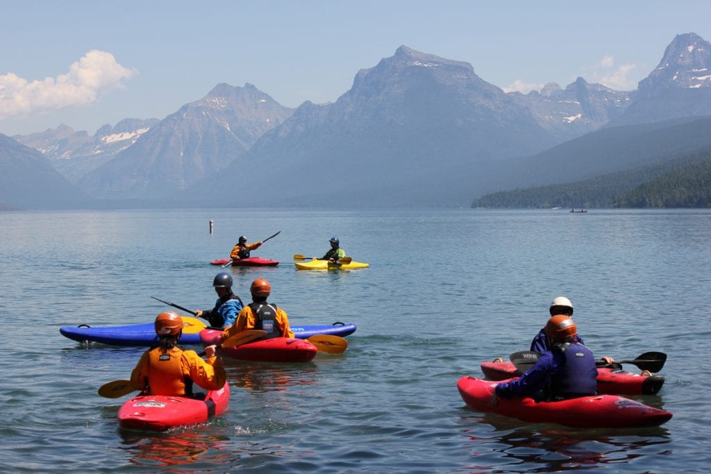 Kayakers in red and yellow kayaks paddle on a lake in Montana with the peaks of Glacier National Park in the backround.