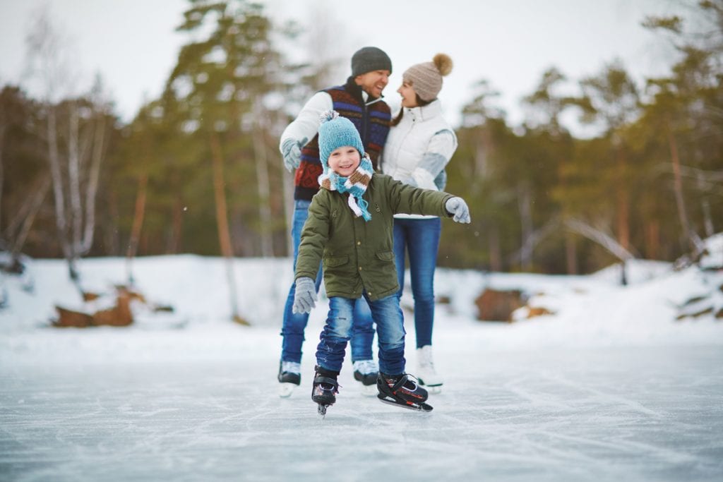 A boy ice skates on a pond in the mountains. He's grinning and his parents are smiling in the background.