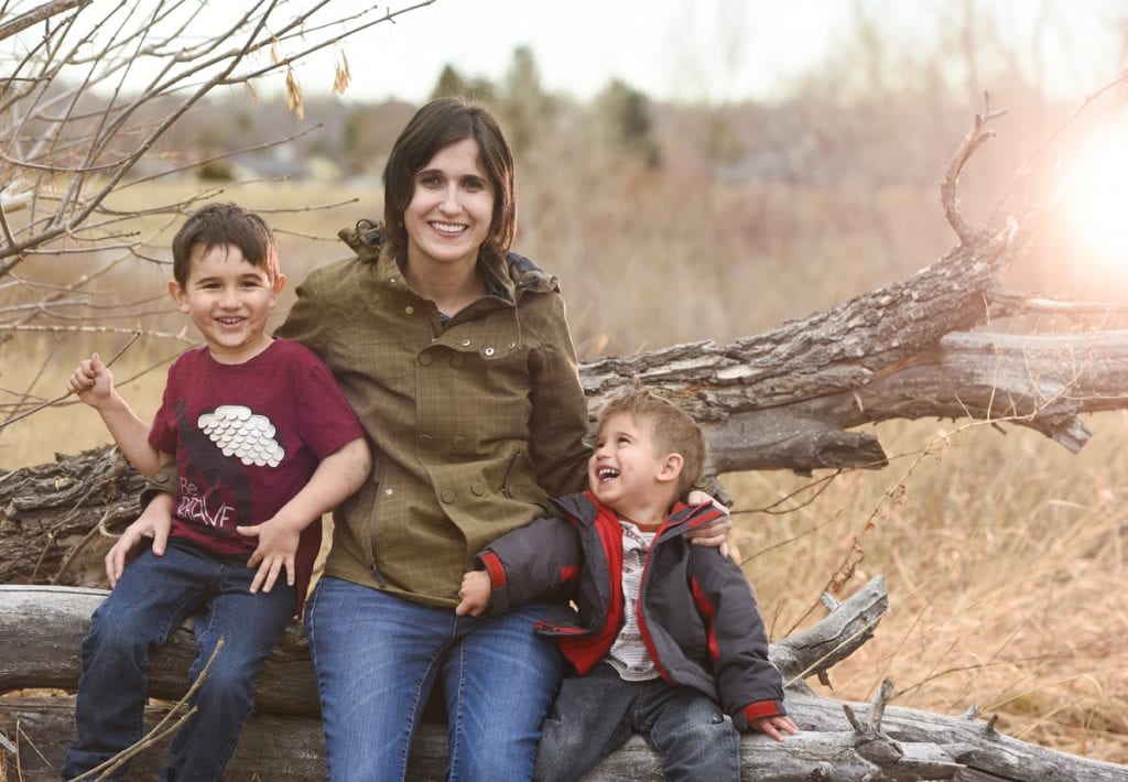 A mom sits on a log and poses with her two young sons, ages 4 and 2. Both kids are laughing and the younger one is looking up at his mom.