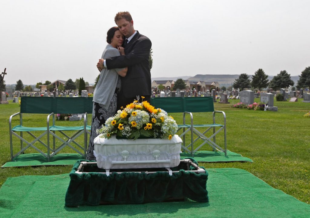 Parents at a funeral for a baby. They hug as a small white casket sits in front of them.
