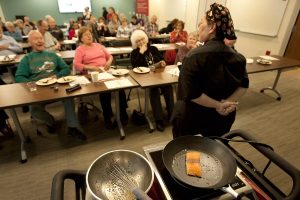 salmon cooks in a skillet while women talks and room of people listen.