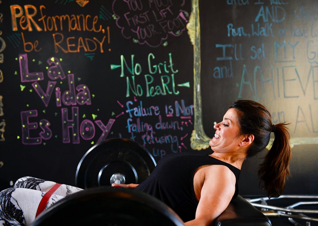 Fighting lymphedema after cancer. Angela lifts a barbell at her gym. Inspirational sayings appear on a wall behind her.