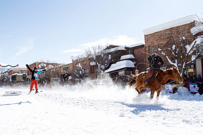 A horse pulls a skier holding a rope down a snow-covered street.
