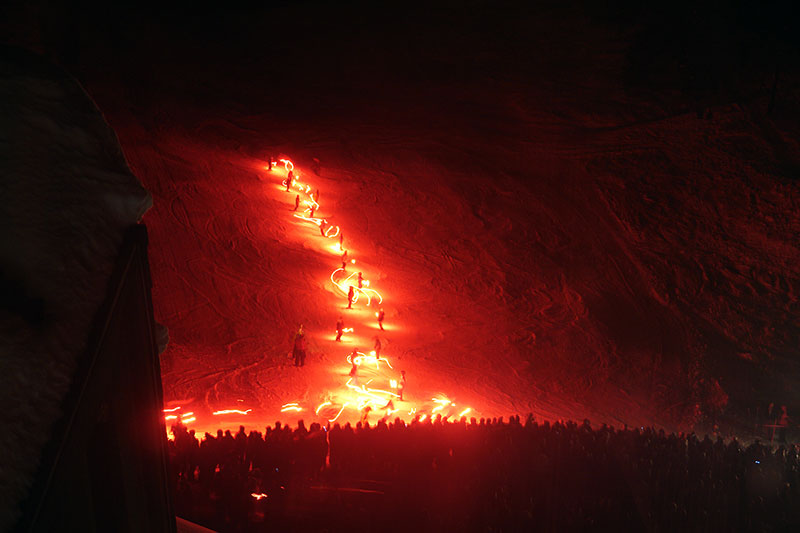 Skiers holding flares ski down a mountain at night in this photo.