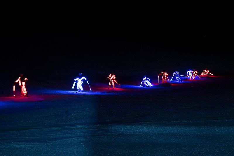 Skiers adorned with LED lights ski at night in this photo.