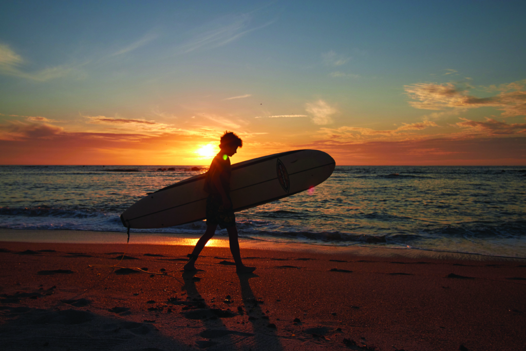 A man walks with a surf board in the sunset