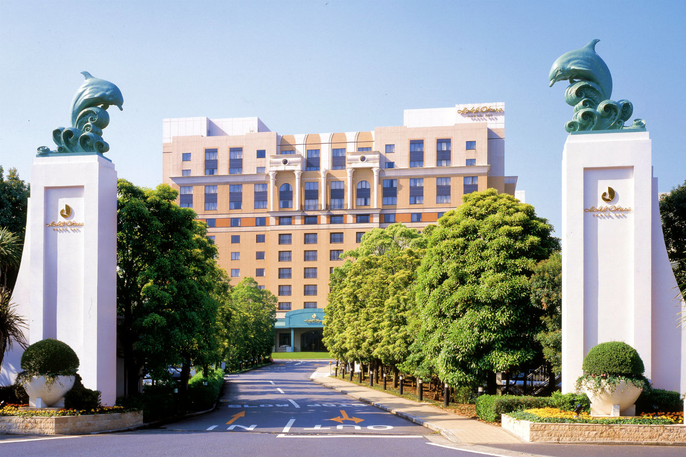 Hotel Okura Tokyo Bay exterior view on a sunny day, framed by white gate pillars topped with dolphin sculptures and surrounded by lush green trees.