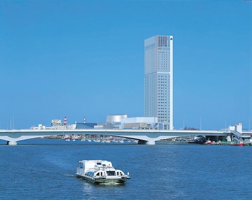 Hotel Nikko Niigata tower overlooking the river with a boat and bridge in the foreground under a clear blue sky.