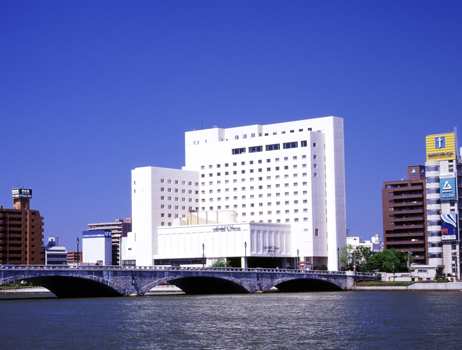 Hotel Okura Niigata, a white mid-rise building beside a river and bridge, set against a vivid blue sky.