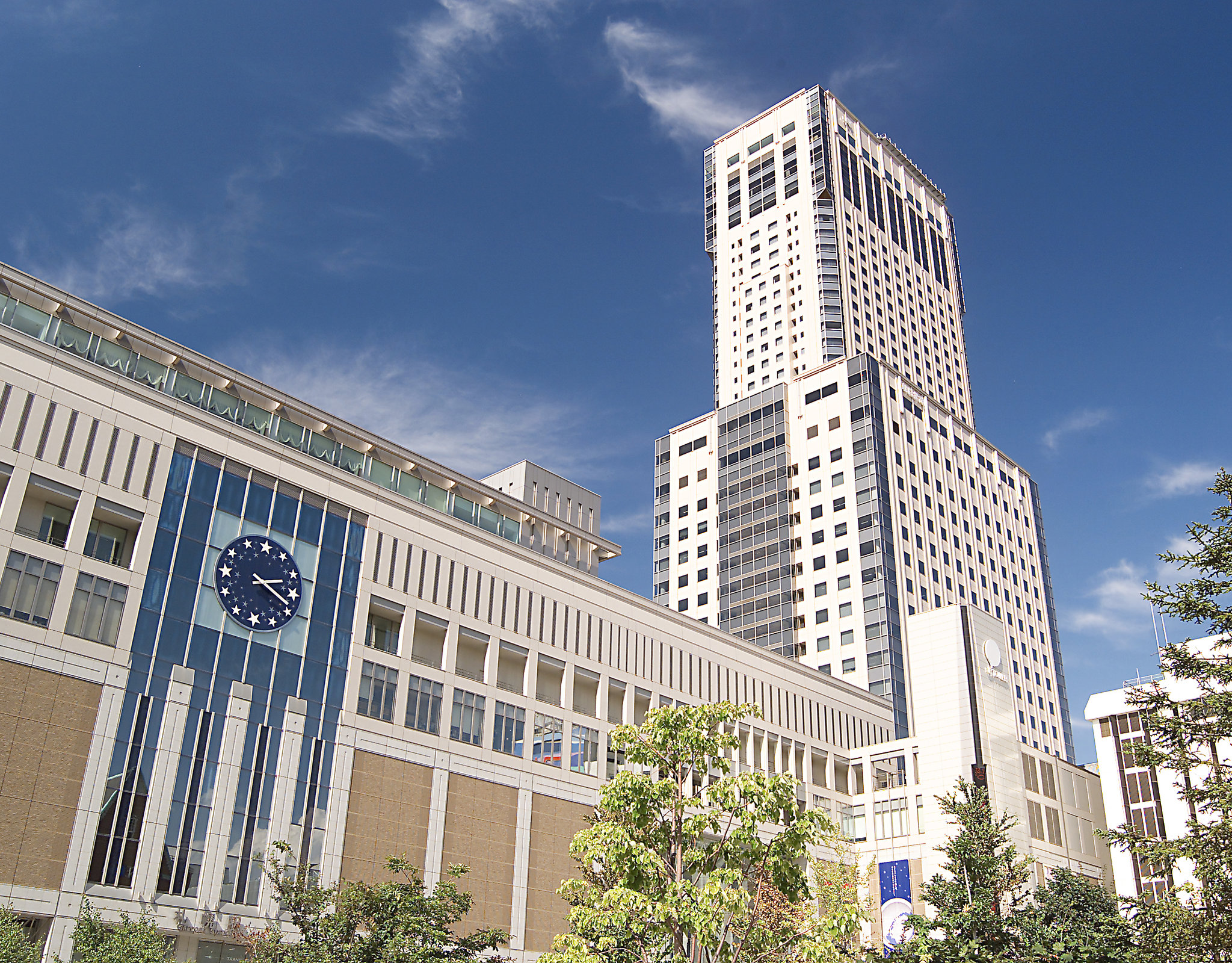 JR Tower Hotel Nikko Sapporo, a striking high-rise building above a modern shopping complex with a large blue clock, set against a vibrant sky.