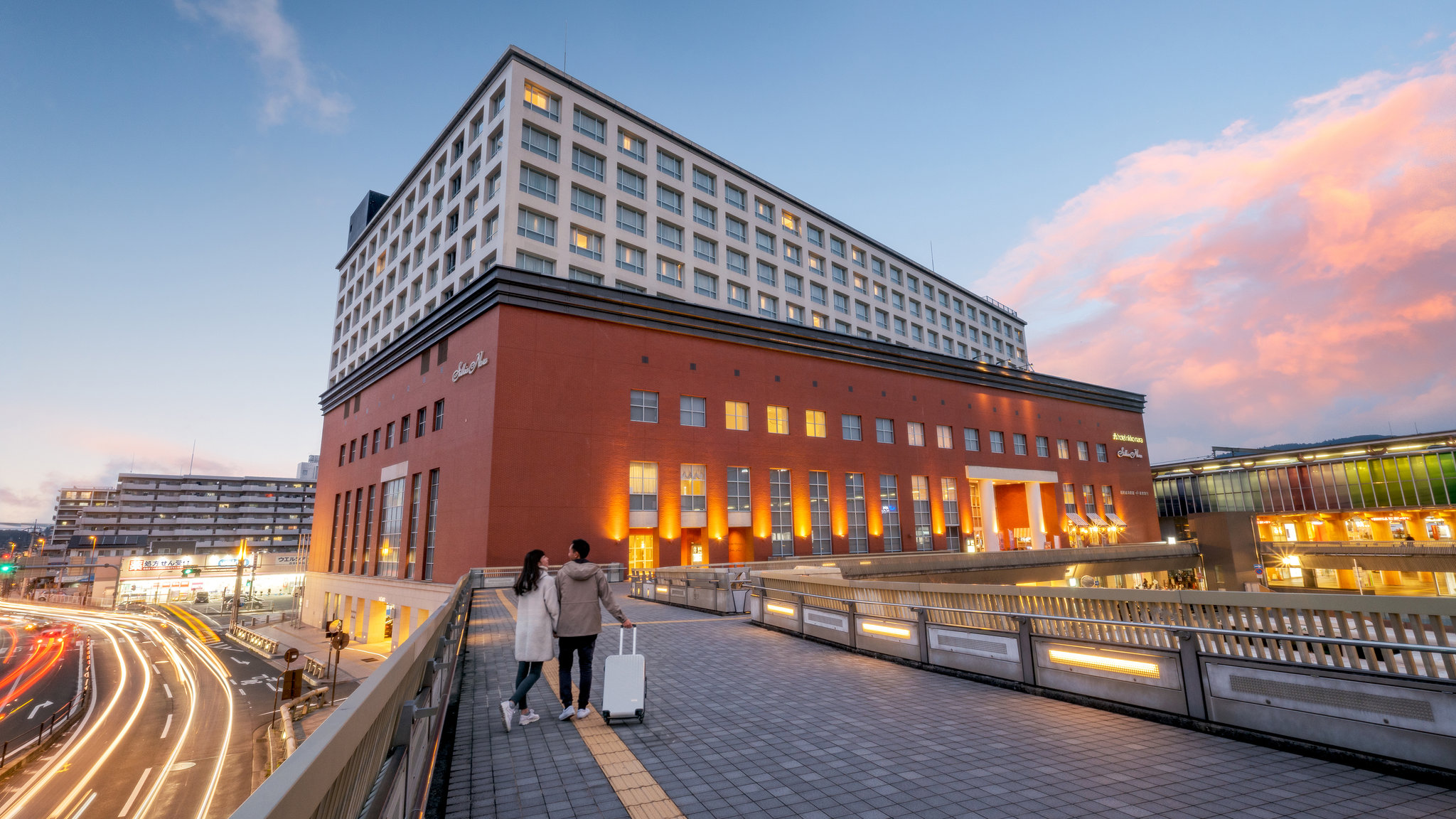 Hotel Nikko Nara at sunset, with warm lighting on its red-brick facade and a couple walking on a pedestrian bridge in the foreground.