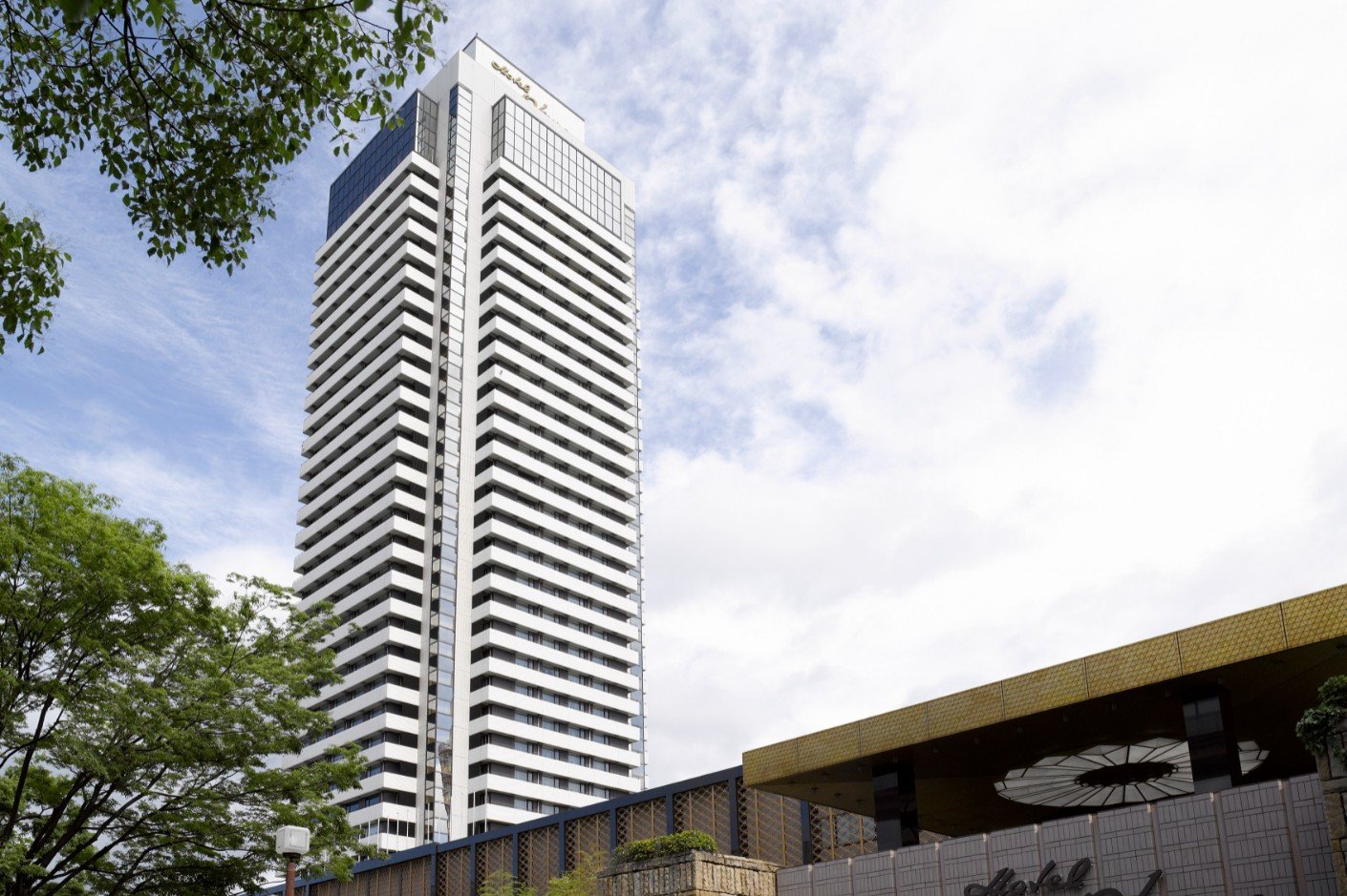 Hotel Okura Kobe, a tall white high-rise with uniform balconies, set beside a modern low-rise structure and framed by trees under a partly cloudy sky.