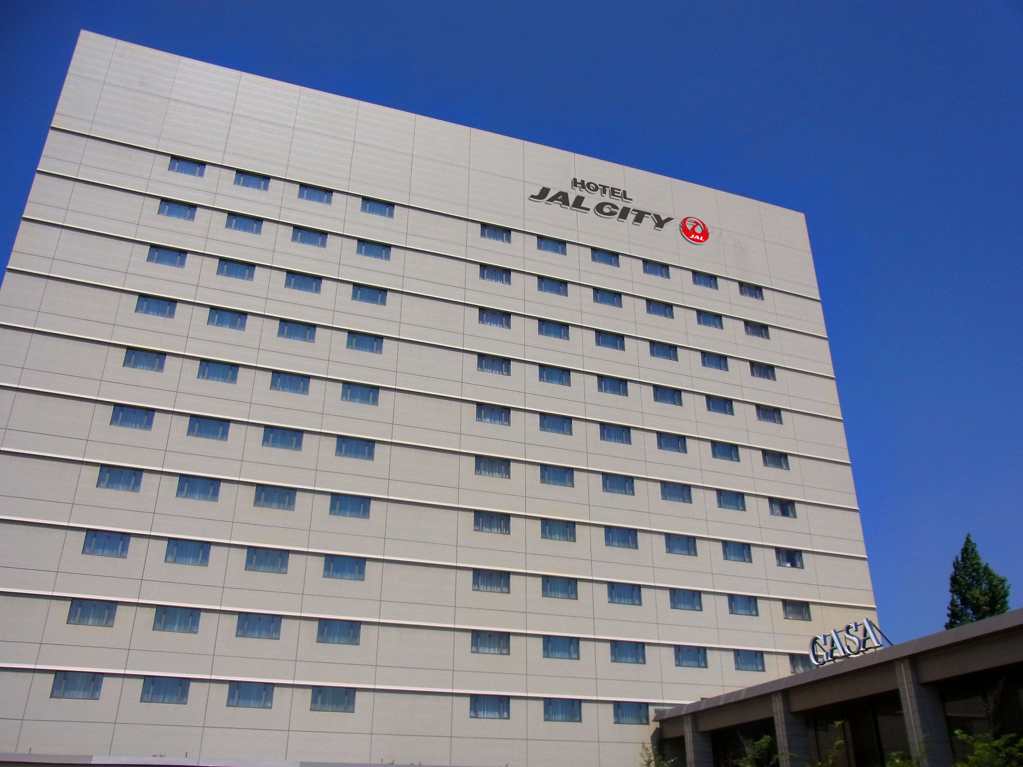 Hotel JAL City Tsukuba, a light-colored mid-rise building with rows of windows and a clear blue sky backdrop.