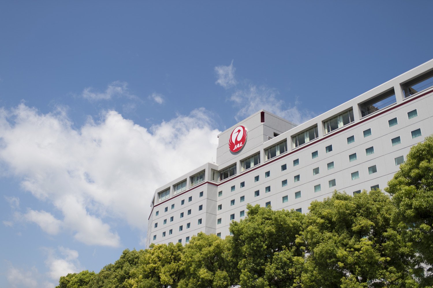 Hotel Nikko Narita, a white modern building with the JAL logo, framed by green trees and a bright blue sky with scattered clouds.
