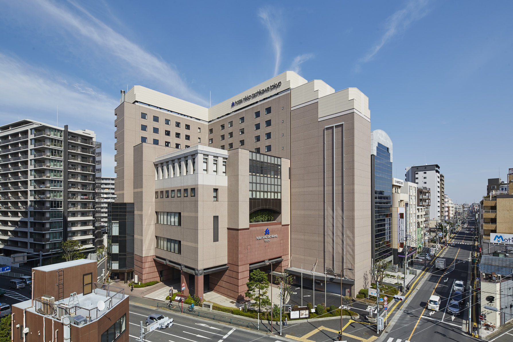 Hotel Nikko Tachikawa Tokyo, a large modern building in a busy urban neighborhood, surrounded by residential and office towers under a clear blue sky.
