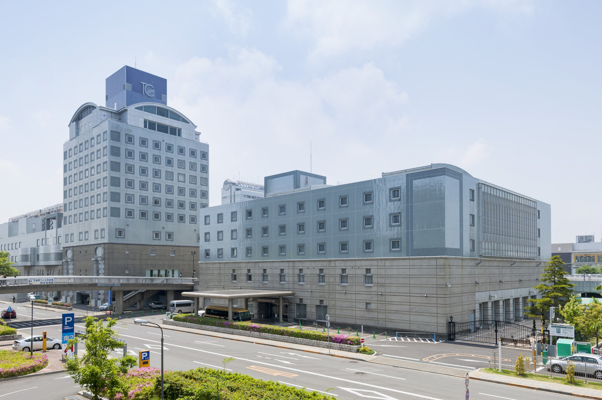 Hotel Nikko Tsukuba, a modern gray-toned building complex near a busy road, featuring a mix of rectangular structures and a connecting skywalk under a hazy blue sky.