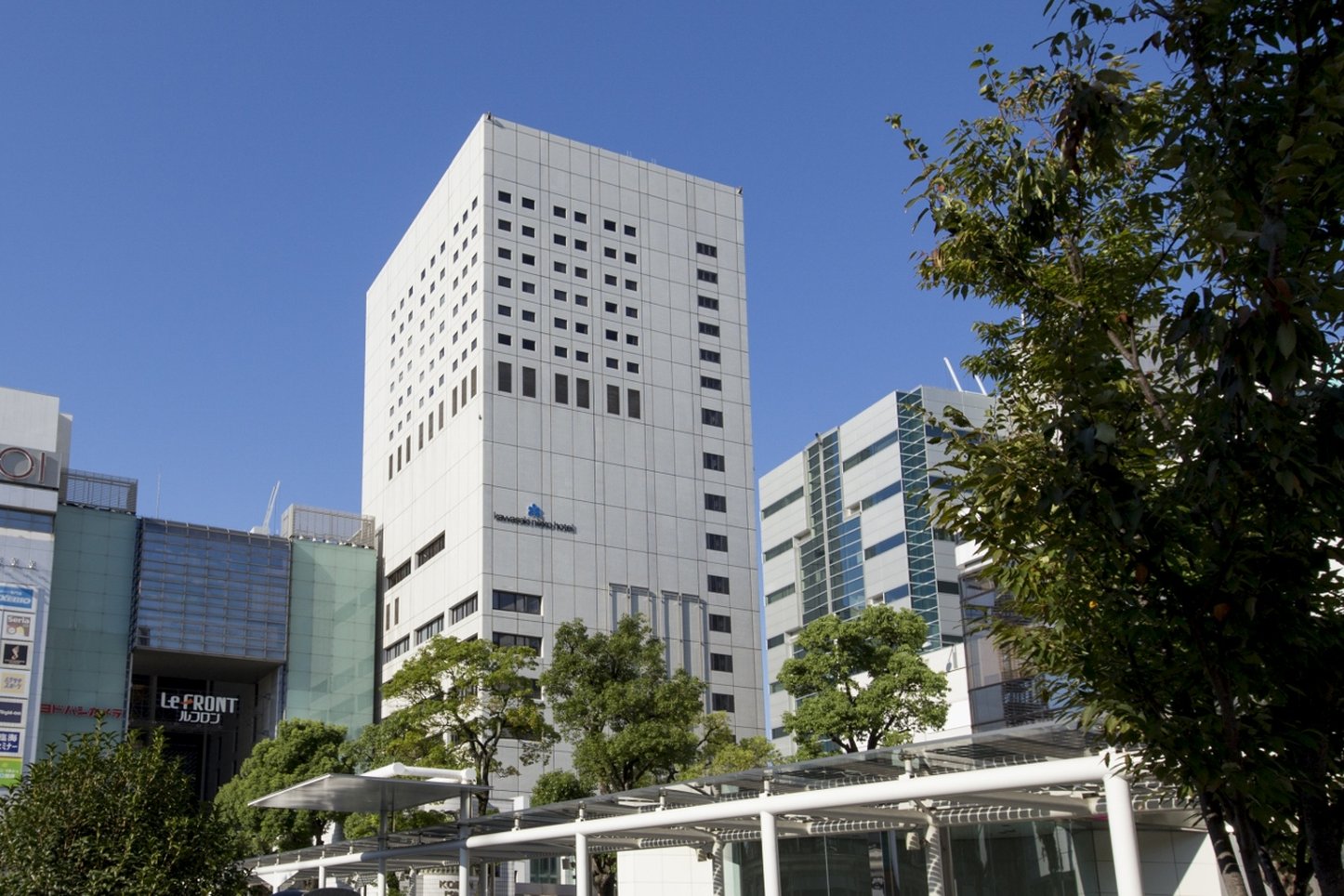 Kawasaki Nikko Hotel, a tall white tower with a grid-like window pattern, situated in a bustling urban area under a clear blue sky.