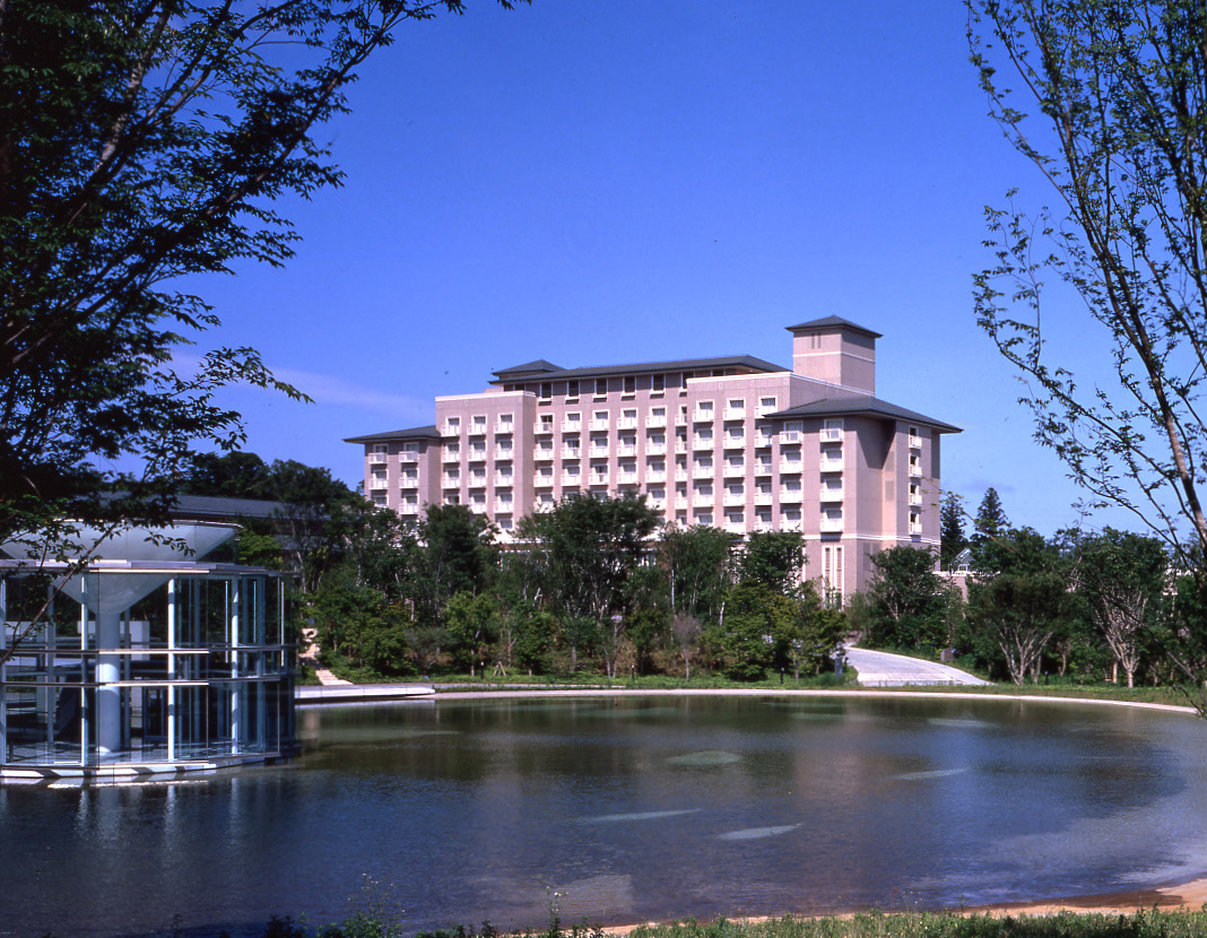 Okura Akademia Park Hotel, a serene mid-rise hotel surrounded by greenery and overlooking a tranquil pond under a bright blue sky.