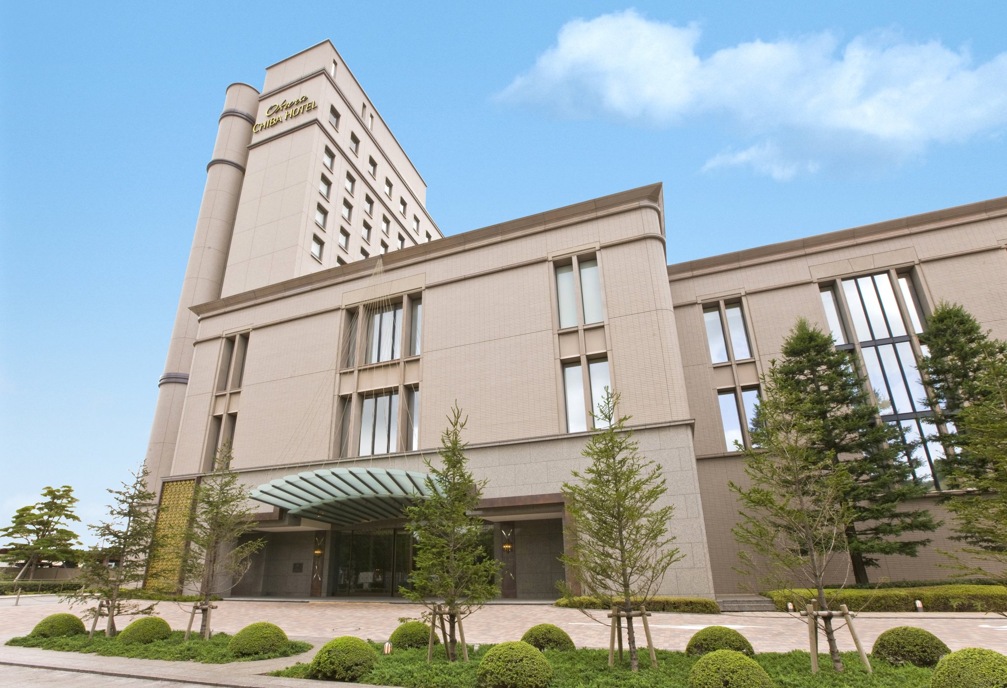 Okura Chiba Hotel, a sophisticated high-rise with a modern beige facade, neatly landscaped greenery, and elegant vertical window accents under a clear blue sky.