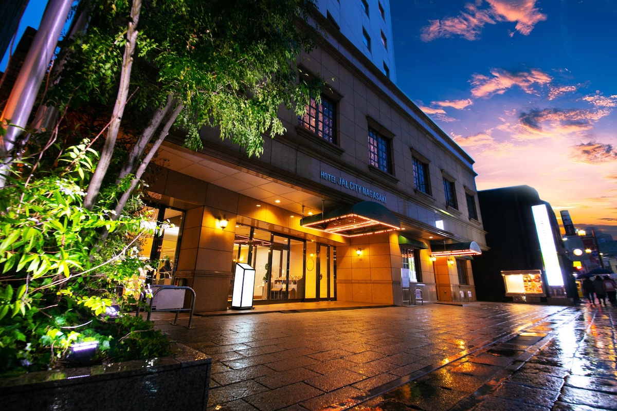 Entrance of Hotel JAL City Nagasaki illuminated at dusk, with glowing signage and reflections on wet pavement under a vibrant sunset sky.