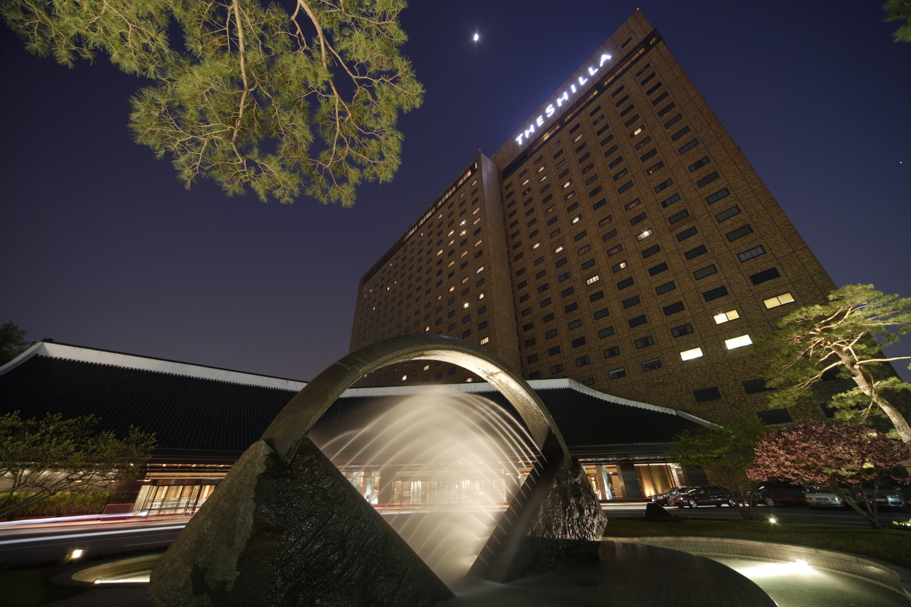 The Shilla Seoul at night, featuring a tall brick facade, illuminated signage, and a modern arch fountain in the foreground.