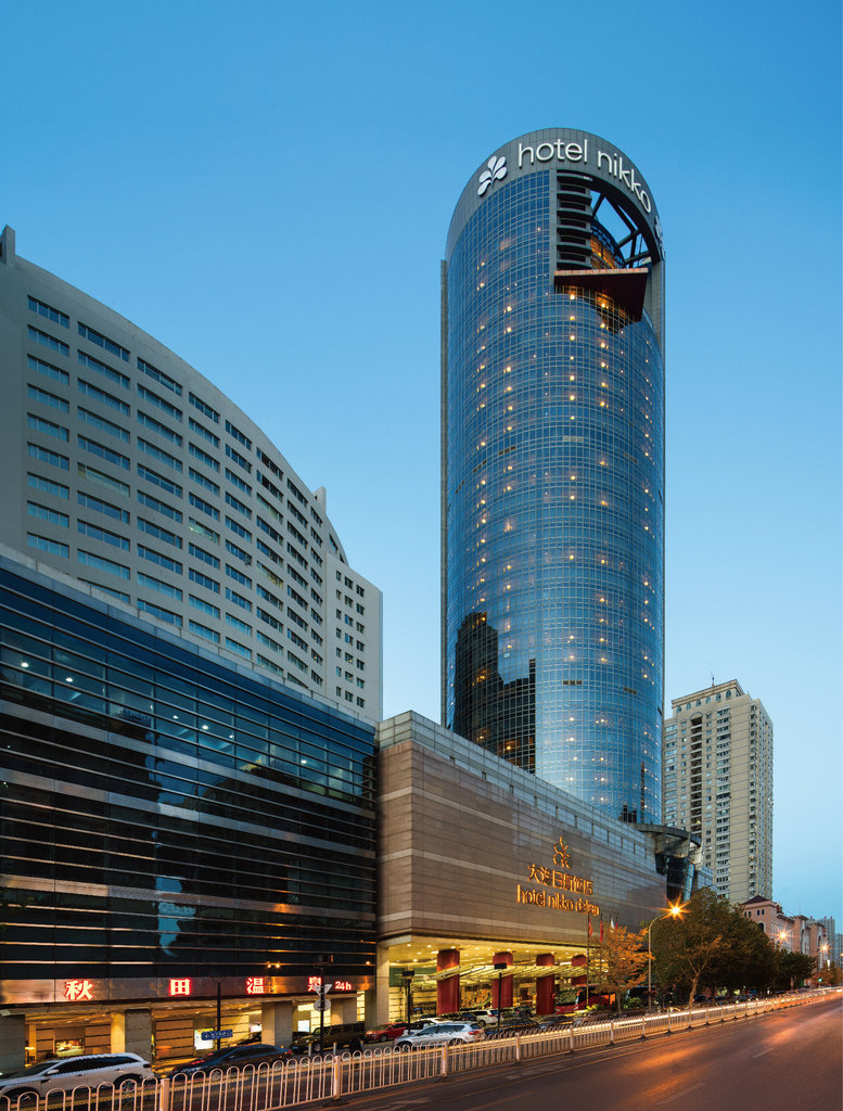 Hotel Nikko Dalian features a sleek cylindrical tower rising above a bustling city street at dusk.