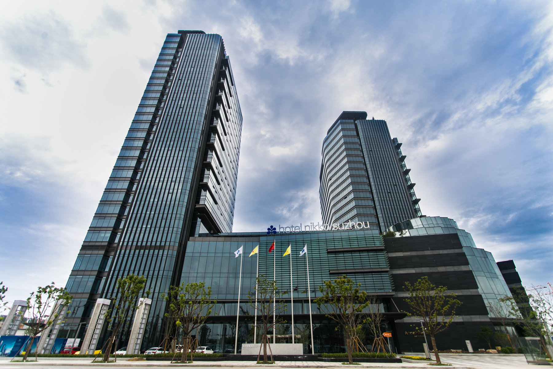 Hotel Nikko Suzhou features two sleek modern towers rising above a glass-fronted entrance, framed by flags and a landscaped plaza.