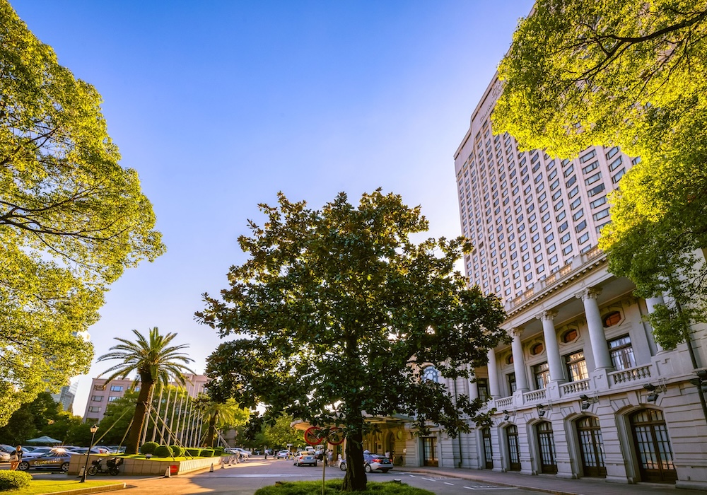 Okura Garden Hotel Shanghai, a grand white historic building with columns and a tall tower, surrounded by lush trees and set against a bright blue sky.